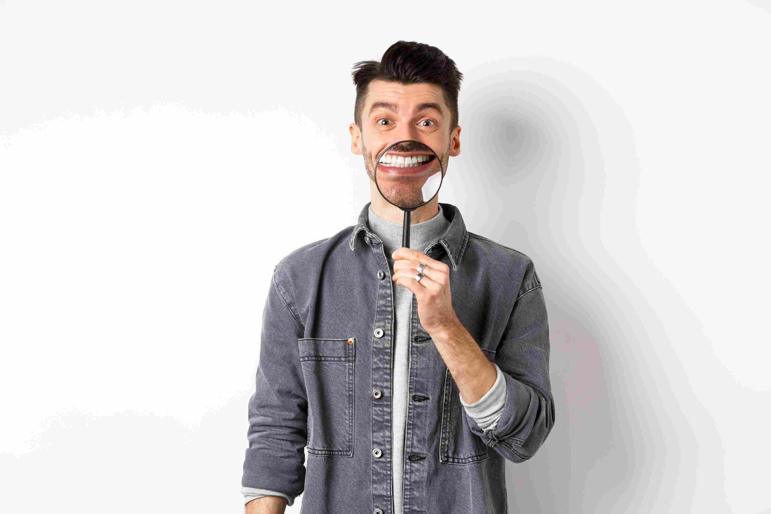 Handsome positive guy showing white perfect smile with magnifying glass, standing against white background